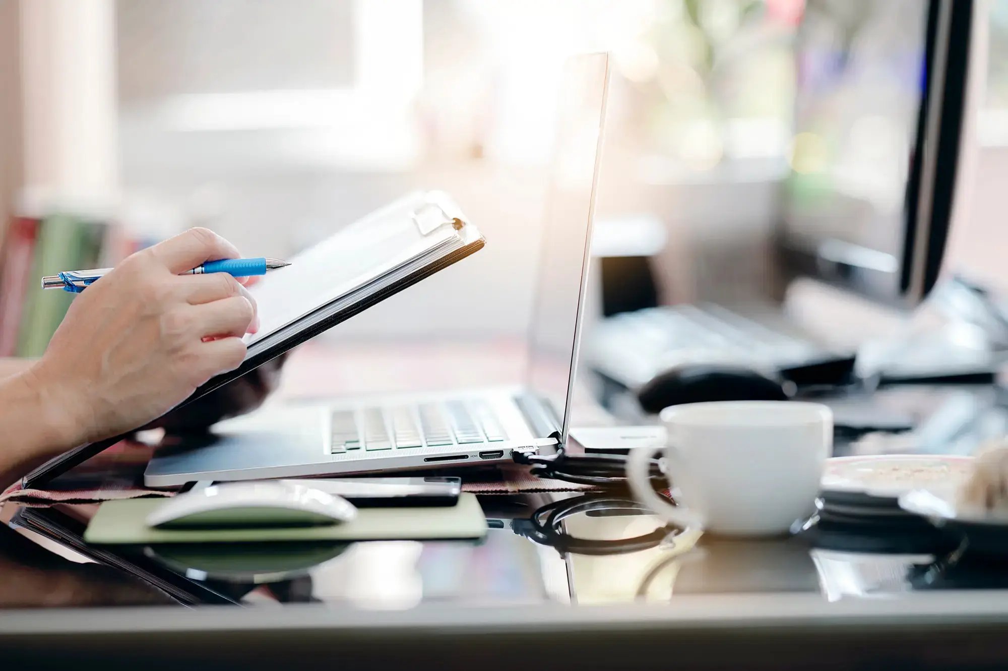 Mano sosteniendo un portapapeles frente a una computadora portátil, en un escritorio con taza de café, gafas y artículos de oficina.