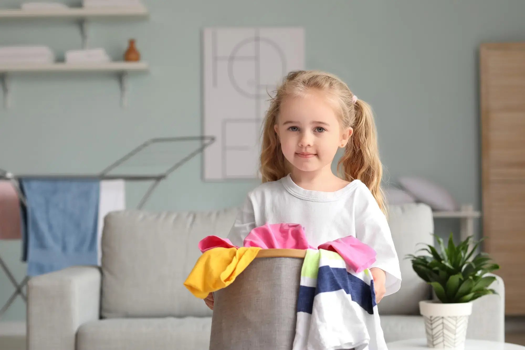 Niña rubia con coletas sosteniendo una canasta llena de ropa en una sala, ayudando con las tareas del hogar.