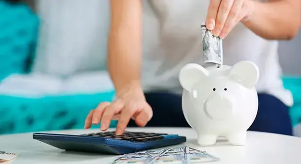 Persona insertando billetes en una alcancía de cerdito mientras usa una calculadora sobre una mesa con dinero, simbolizando ahorro y control financiero.