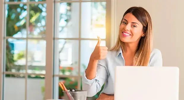 Mujer sonriendo frente a una computadora portátil, levantando el pulgar en señal de aprobación en un entorno de oficina con luz natural.