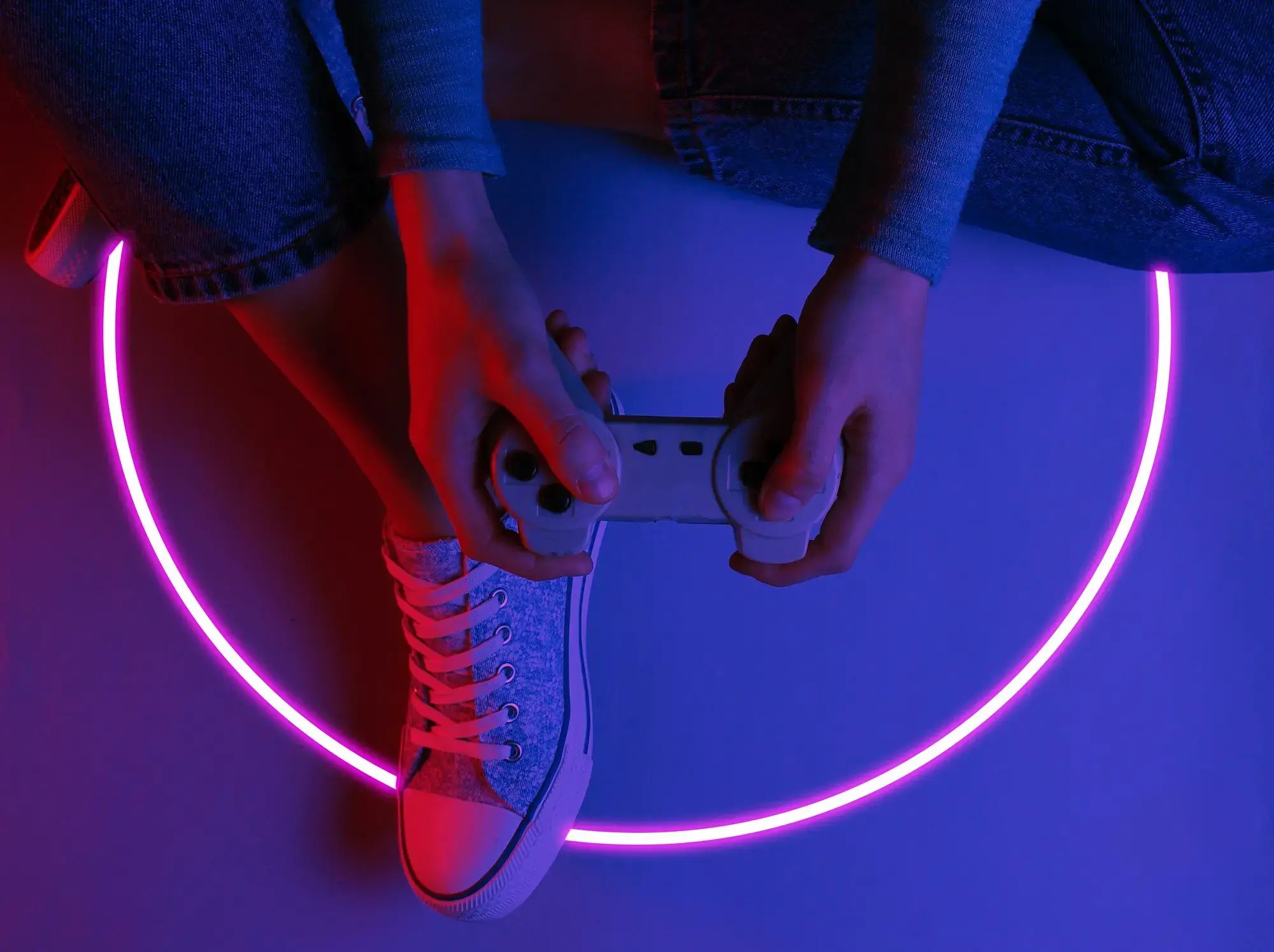 Persona con control de videojuego sentada en el suelo, iluminada con luces de neón rosa y azul en ambiente oscuro.