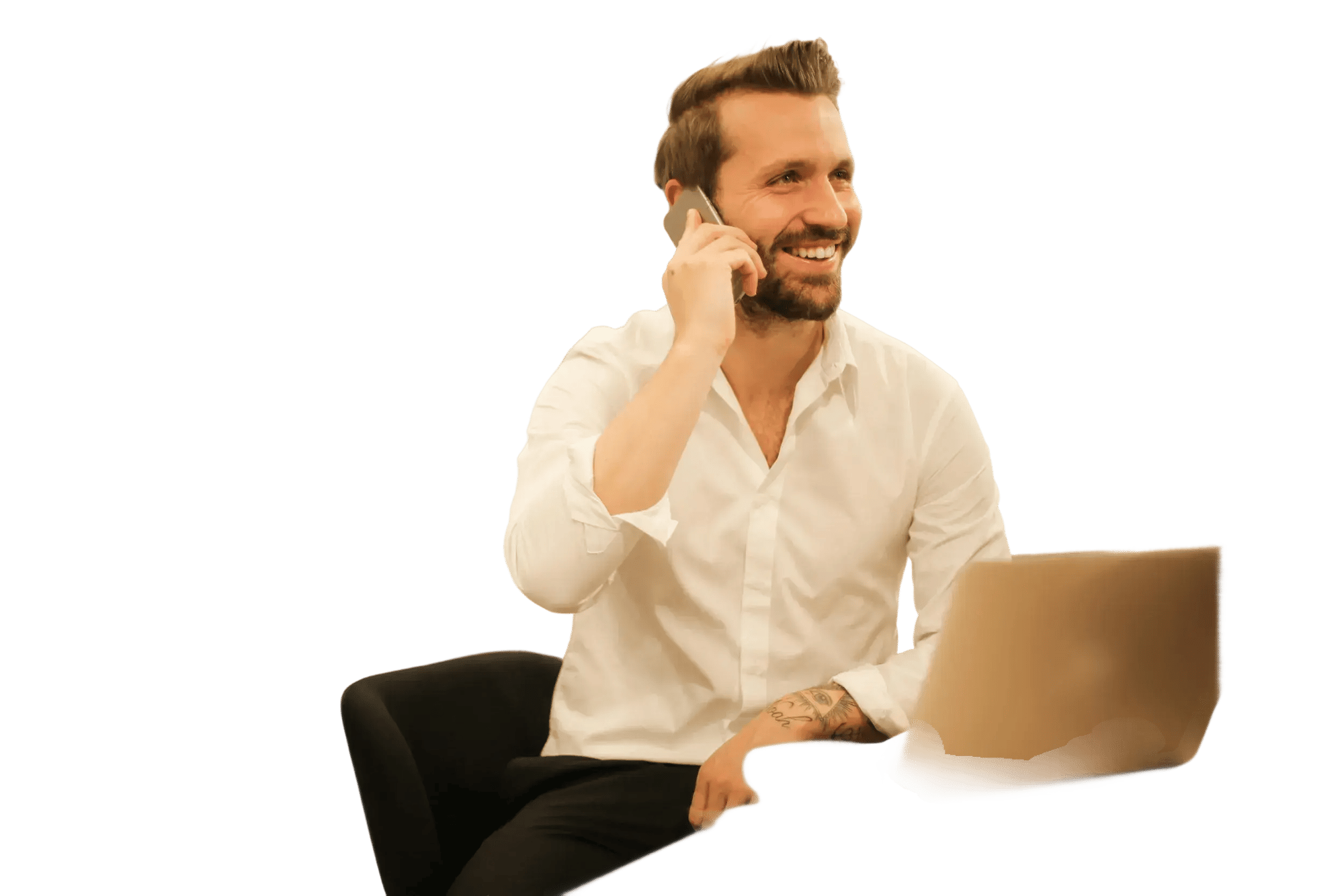 Hombre sentado con camisa blanca hablando por teléfono mientras usa una computadora portátil.