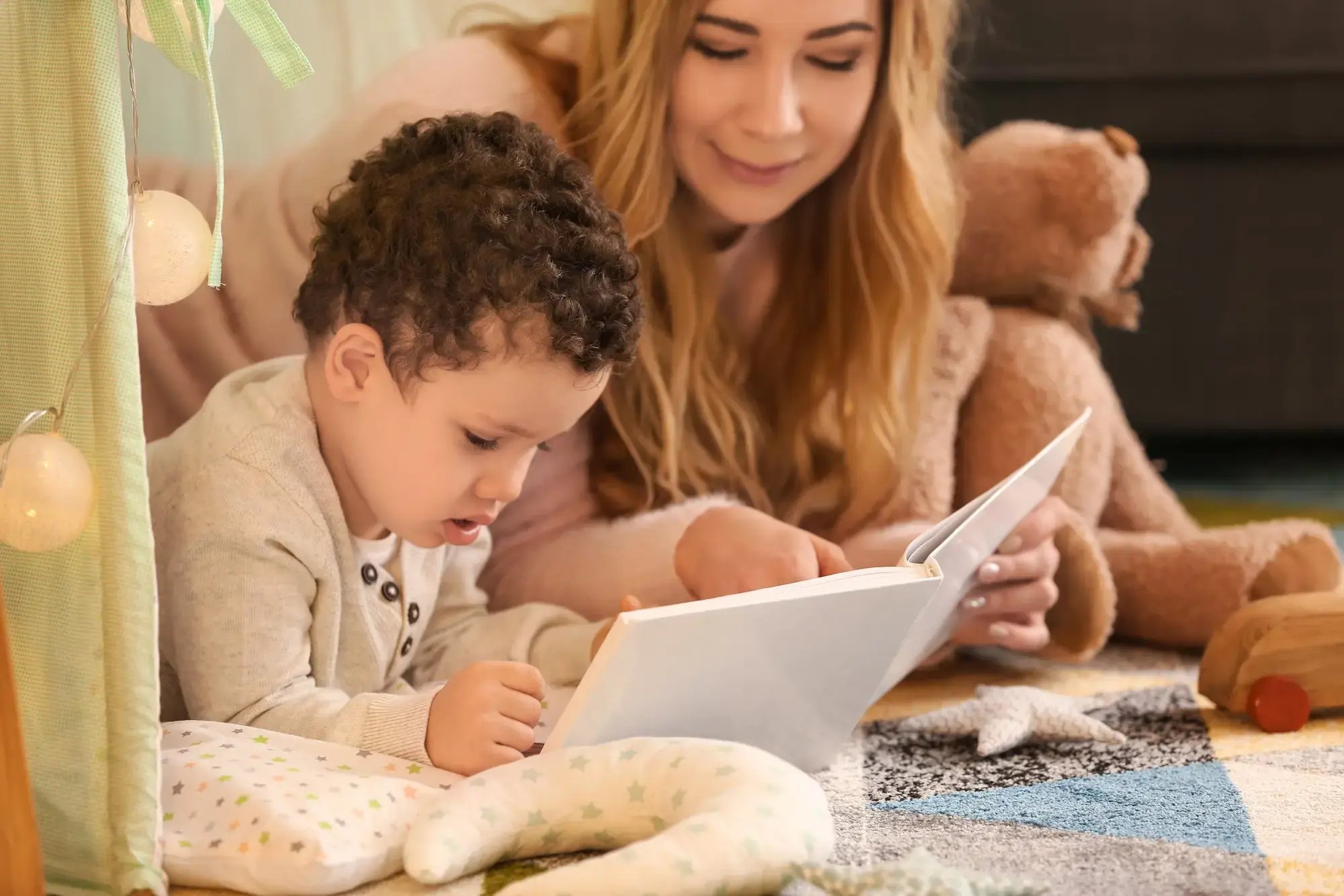 Niño pequeño leyendo un libro acompañado por una mujer, sobre una alfombra con cojines y juguetes de peluche.