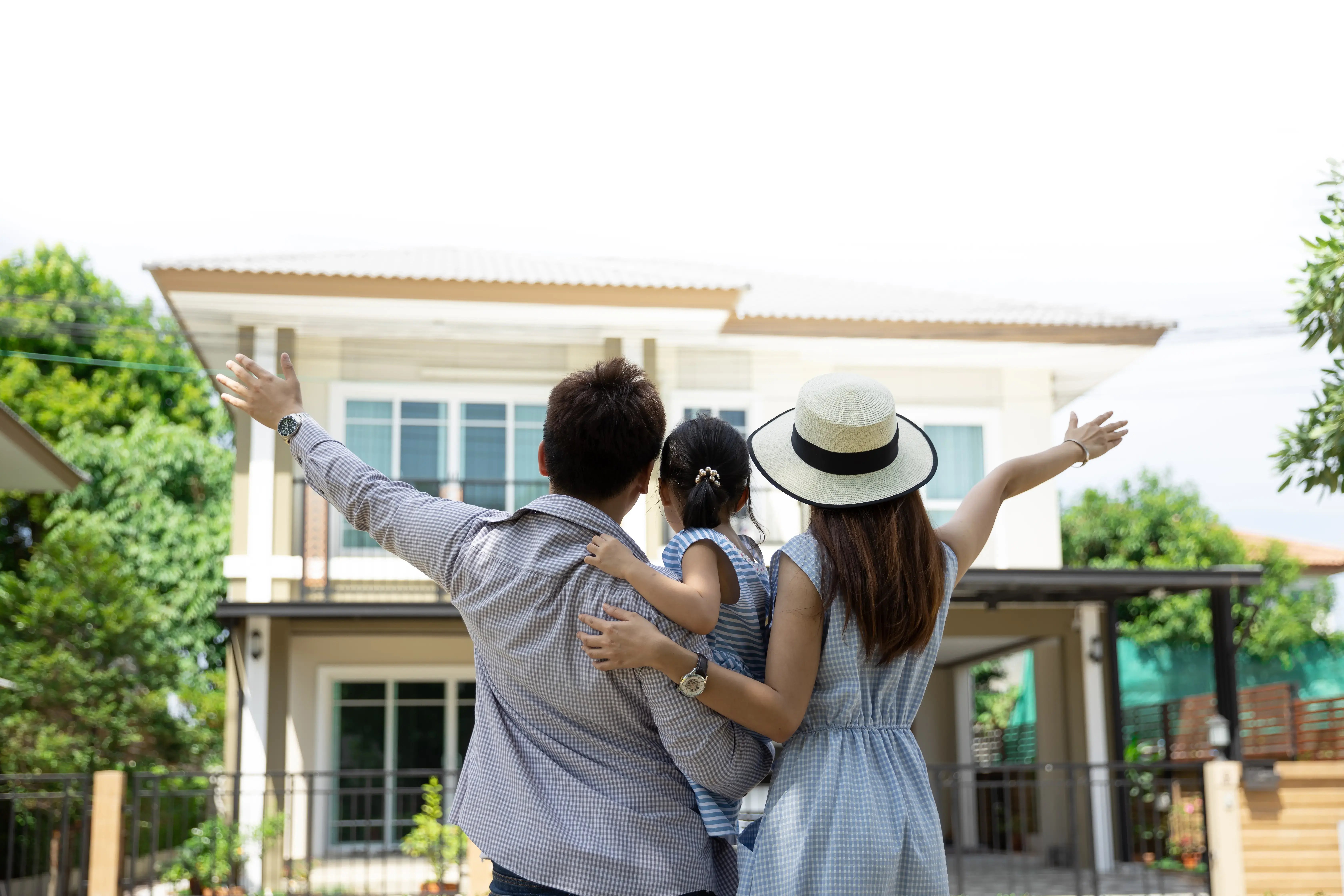 Familia compuesta por padre, madre e hija observando con alegría su nuevo hogar desde el exterior, con los brazos extendidos en señal de emoción.