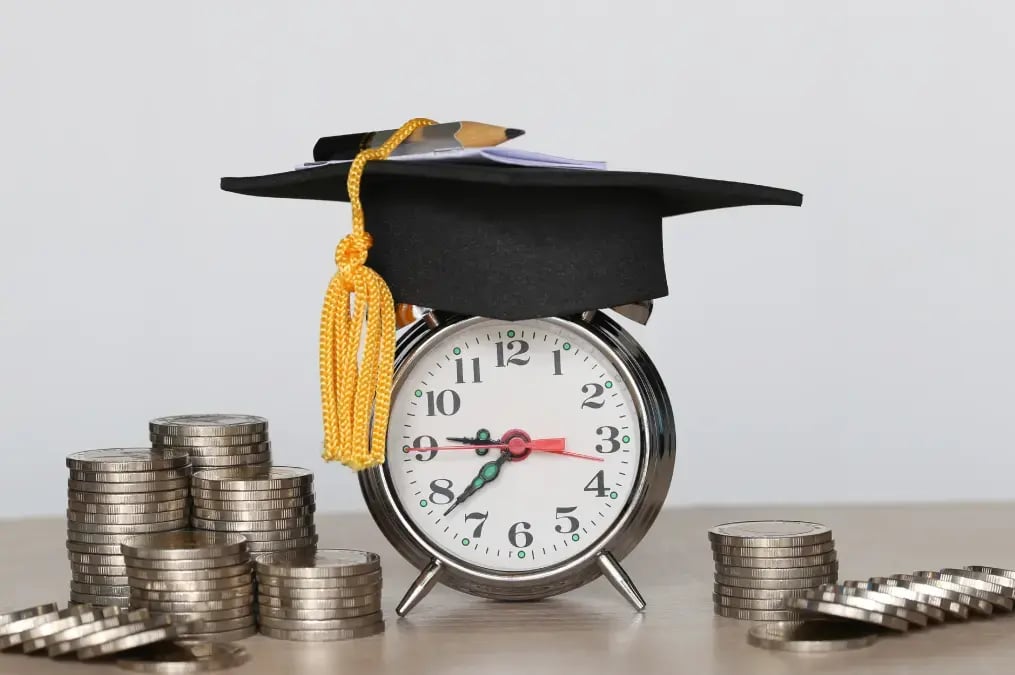Reloj despertador con birrete de graduación y monedas apiladas alrededor.