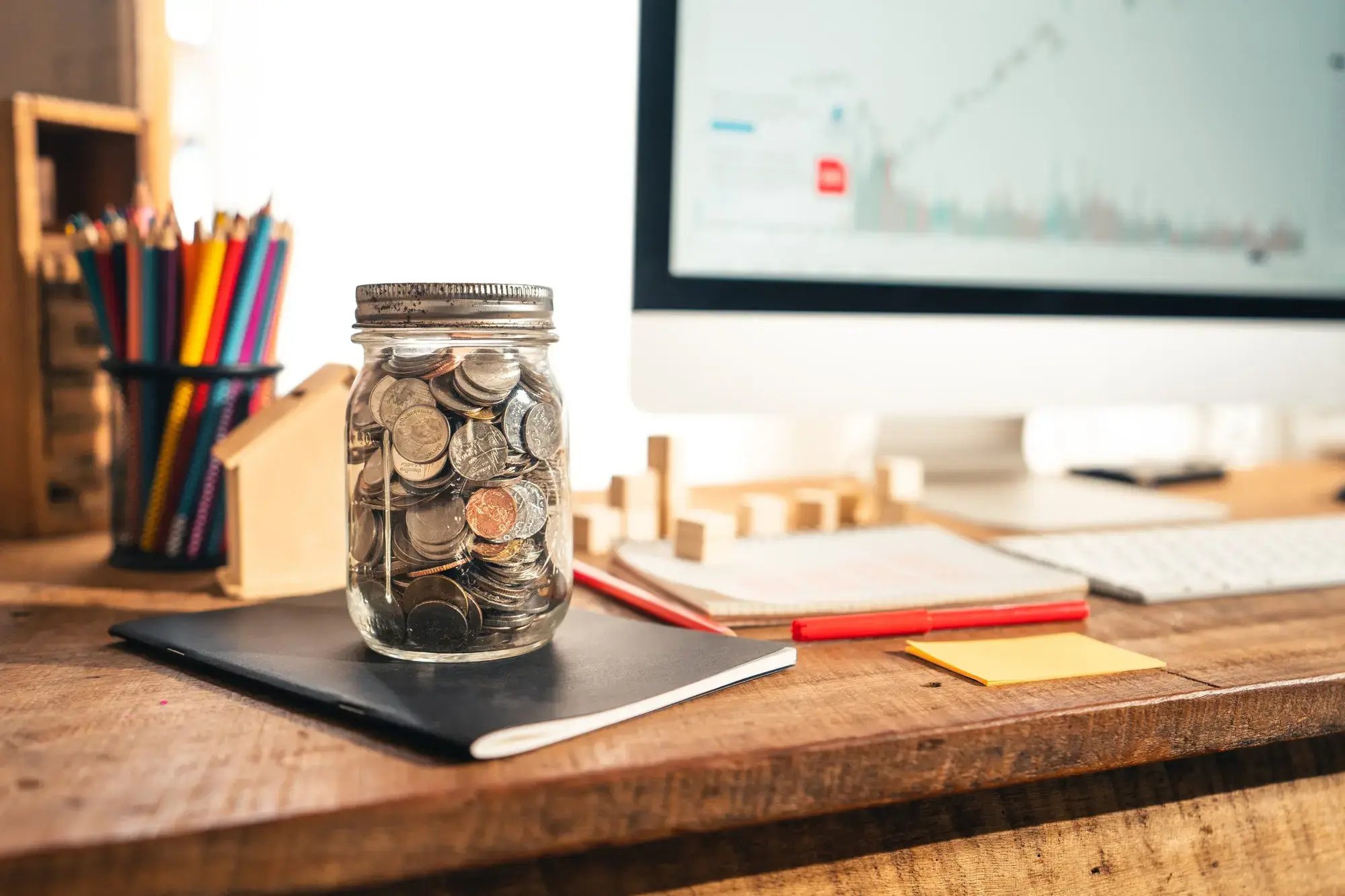 Frasco lleno de monedas sobre escritorio con computadora y gráficos financieros, Banco Nacional de Costa Rica BNCR.