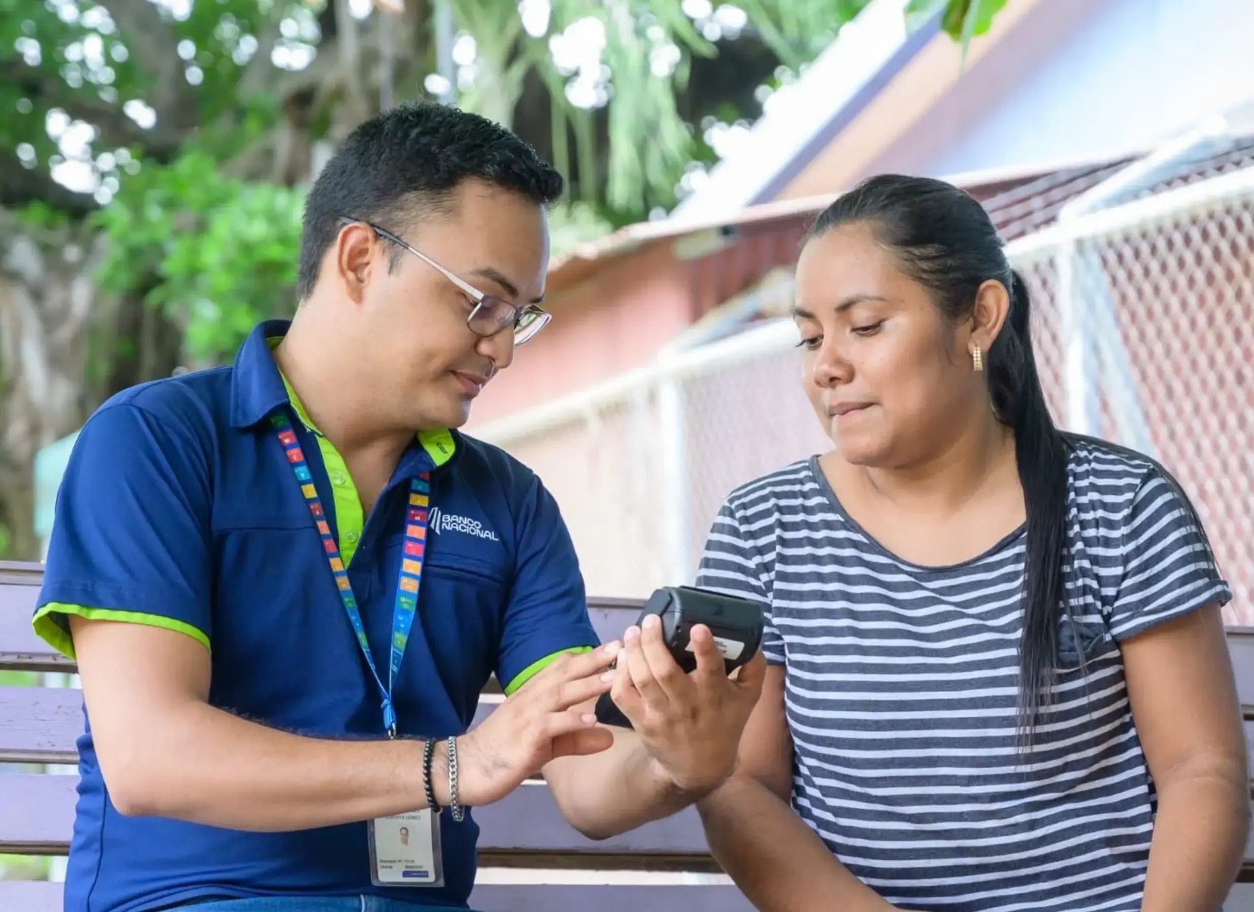 Asesor del Banco Nacional muestra información a una clienta en un dispositivo electrónico al aire libre.