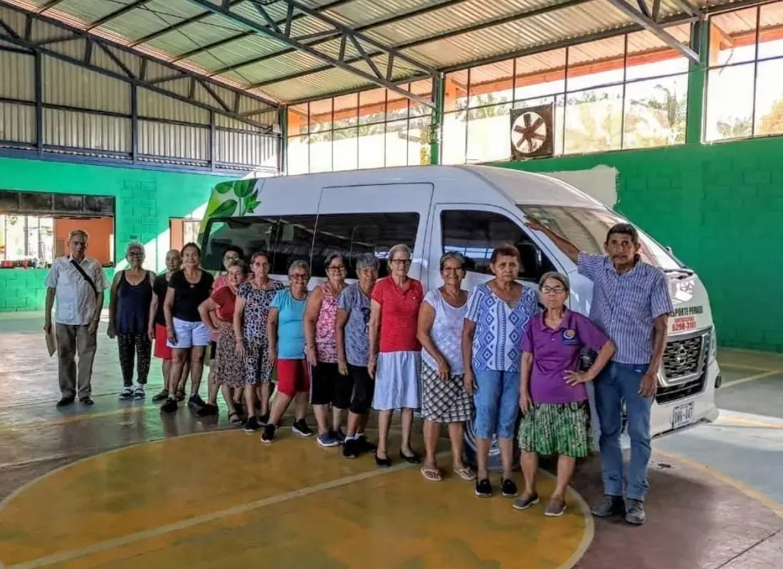 Grupo de adultos mayores en fila frente a un microbús blanco con diseño verde, dentro de un gimnasio techado.