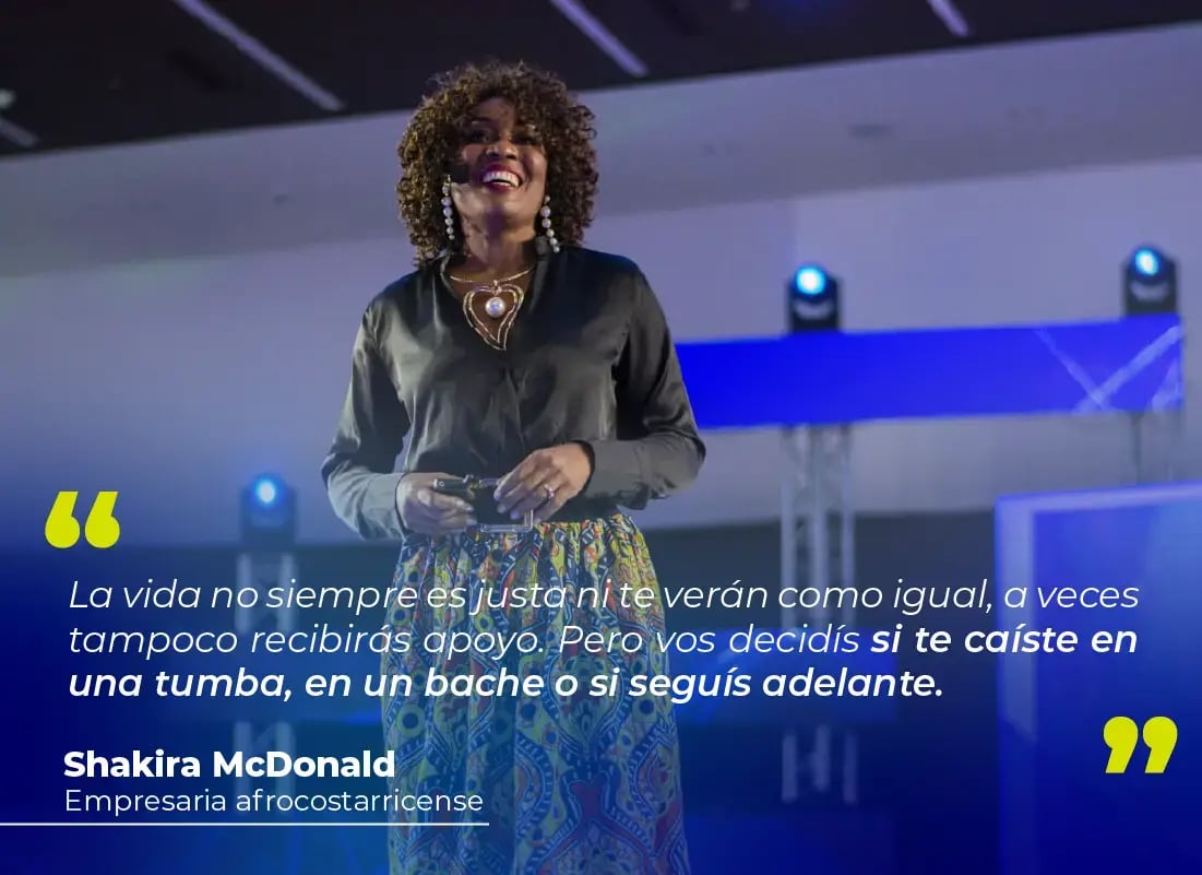 Empresaria afrodescendiente dando una charla motivacional sobre el escenario con iluminación azul y fondo con pantallas.