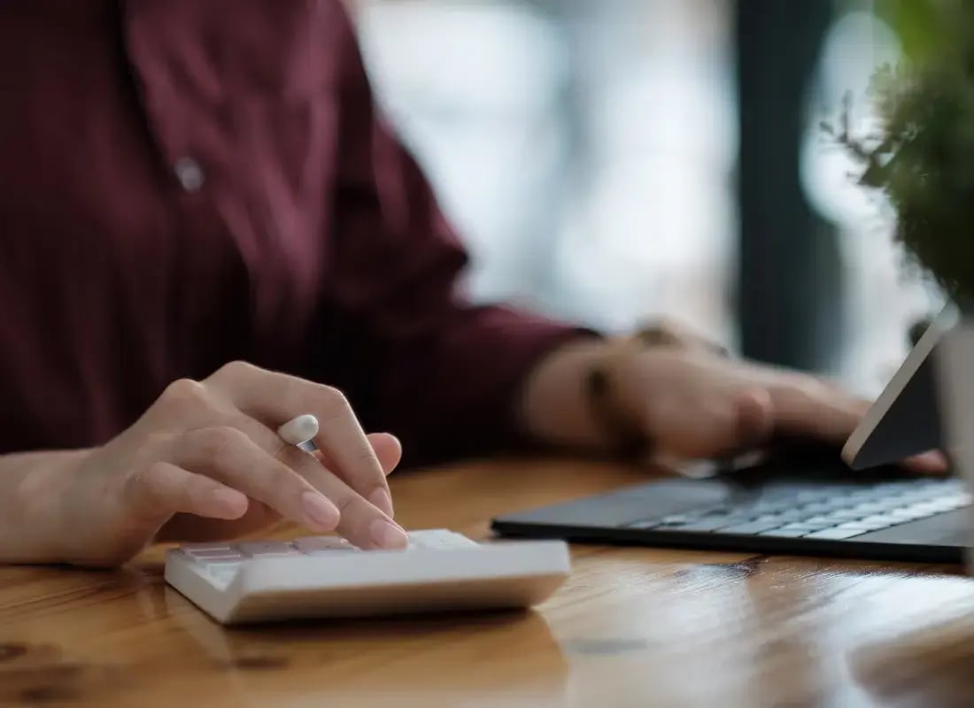 Persona usando calculadora blanca mientras trabaja en una laptop en un escritorio de madera.