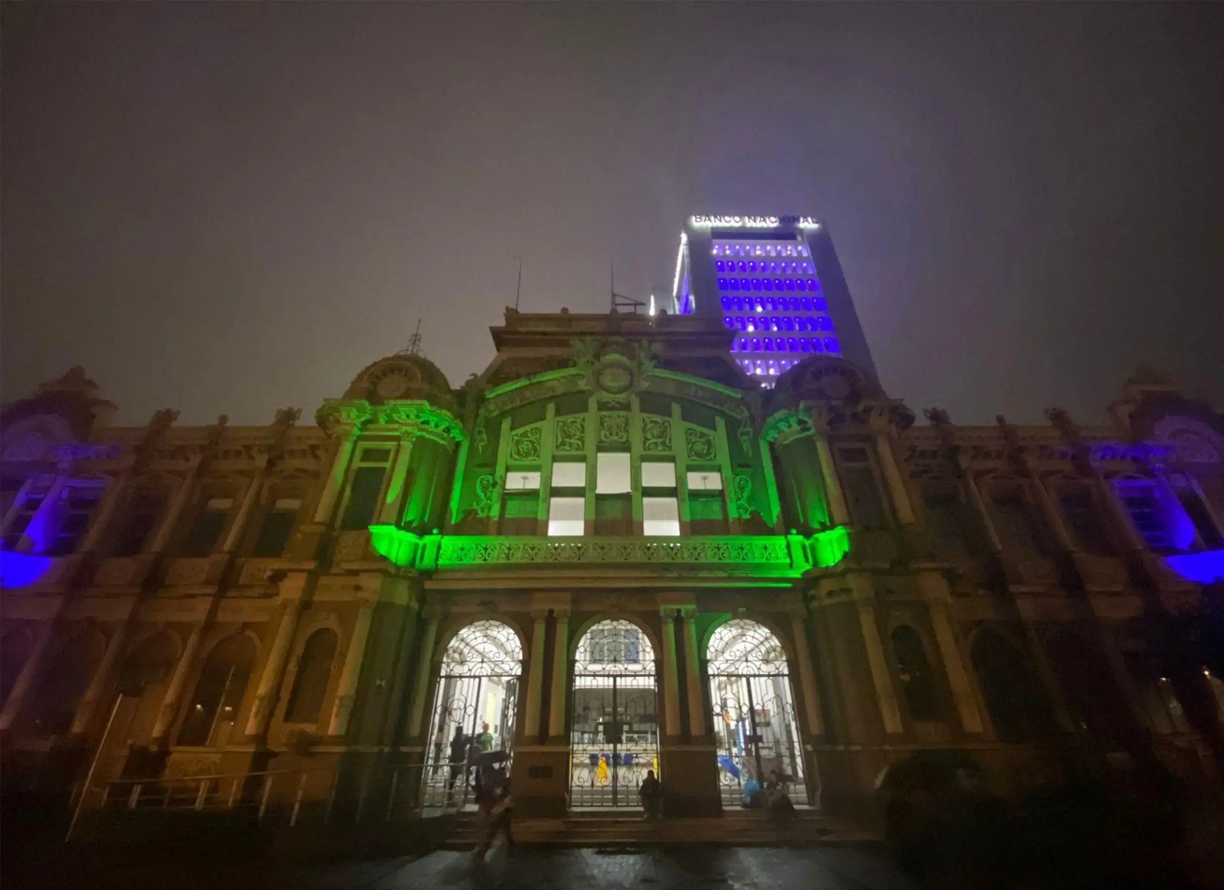 Fachada del edificio histórico del Banco Nacional de Costa Rica iluminada con luces verdes y azules durante la noche.