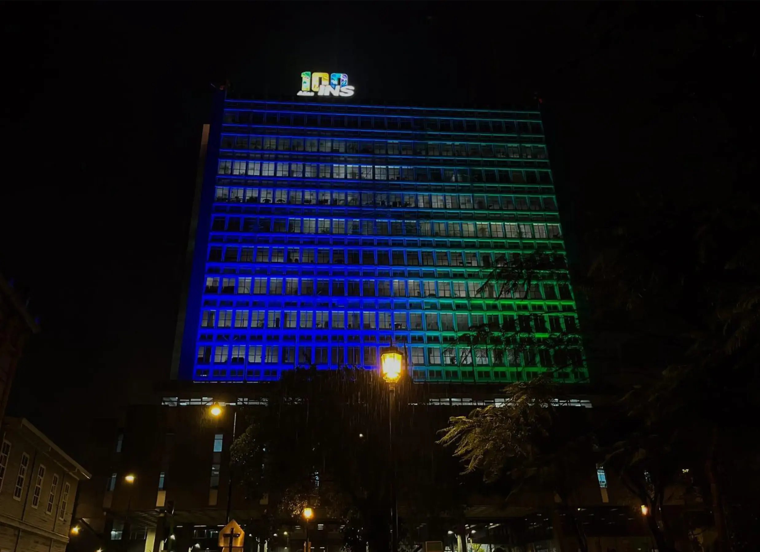 Edificio del Instituto Nacional de Seguros iluminado de azul y verde durante la noche con su logotipo en la parte superior.