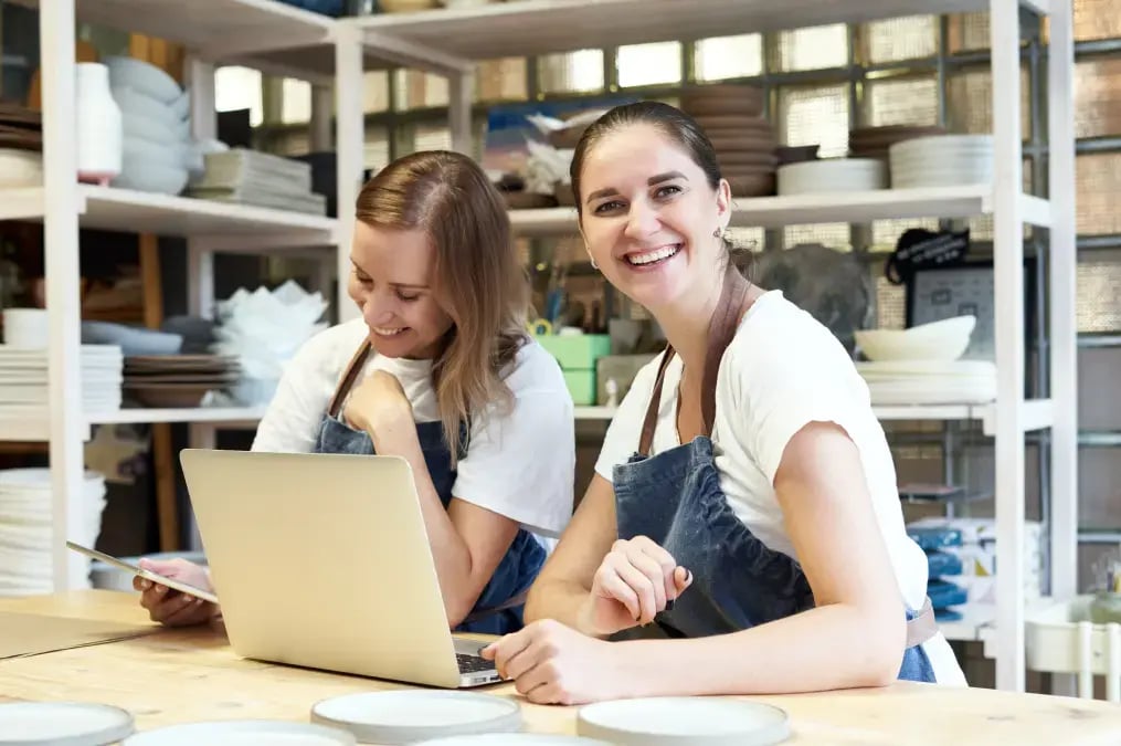 Dos mujeres emprendedoras con delantal trabajando en su negocio de cerámica con una computadora portátil.