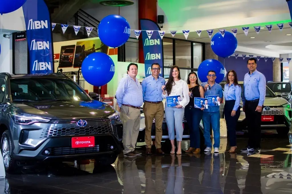 Grupo de personas en agencia Toyota junto a vehículos y globos azules con logotipo del Banco Nacional