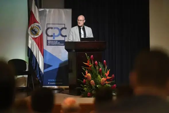Hombre exponiendo en un podio junto a banderas y banner del Consejo de la Competitividad.