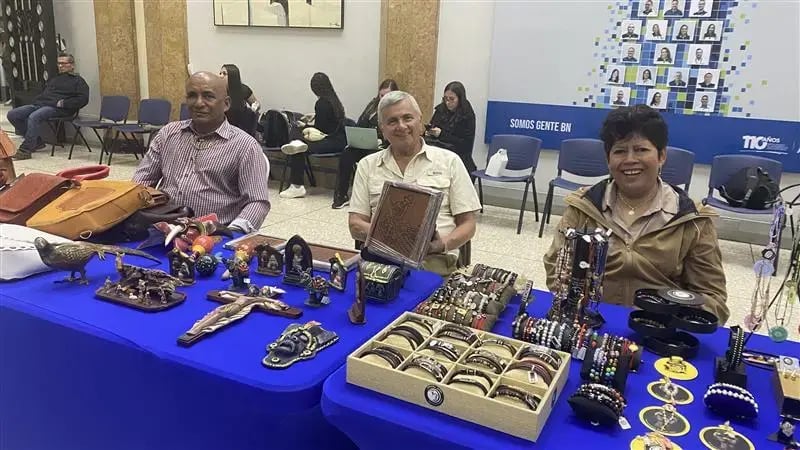 Artesanos exponen joyería, artículos religiosos y productos de cuero en mesas con manteles azules durante la Feria Anual de Artesanos con Discapacidad del Banco Nacional.