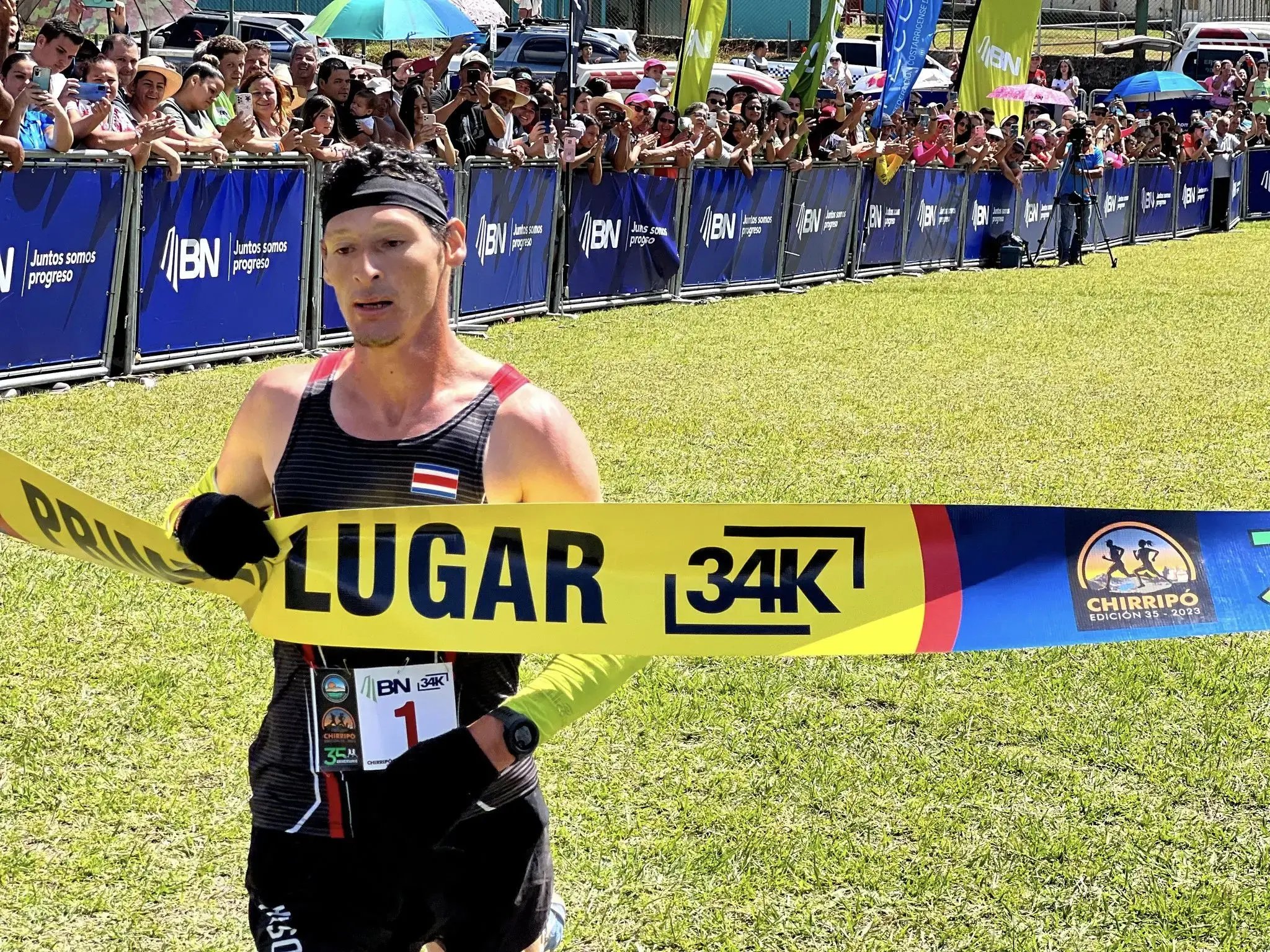 Corredor cruza la meta en primer lugar en la carrera Chirripó 2023, edición 34K, patrocinada por el Banco Nacional, con público animando detrás de las vallas.