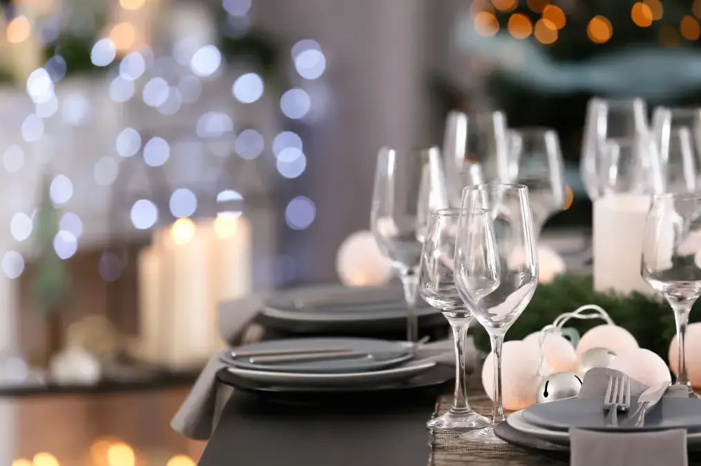 Mesa elegante de Navidad decorada con copas de cristal, velas encendidas y luces festivas desenfocadas al fondo.