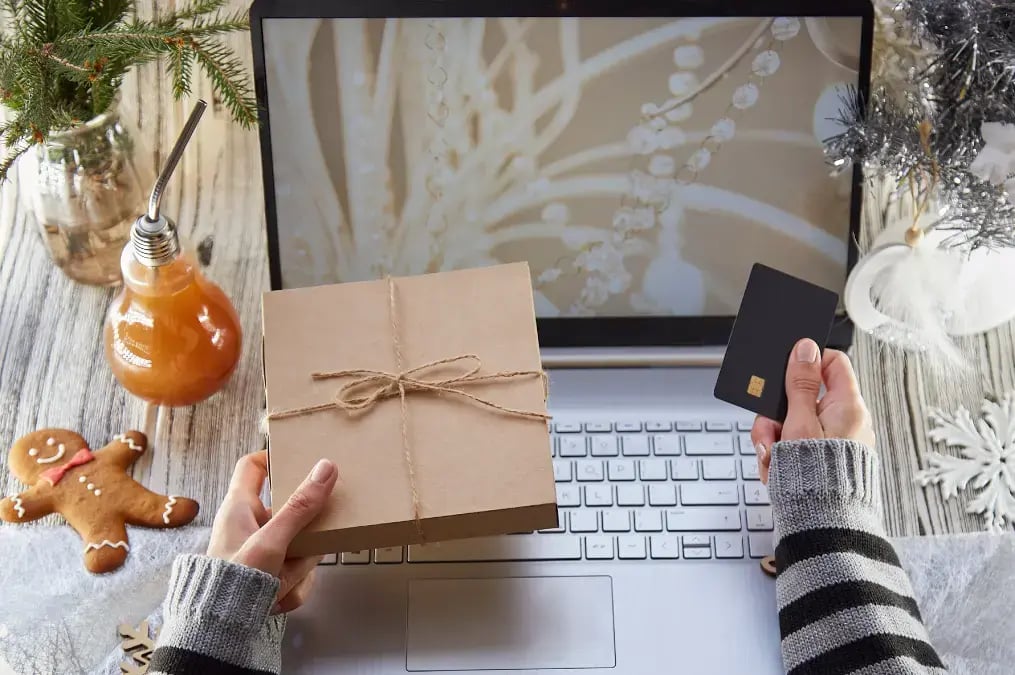 Compra en línea navideña: persona con tarjeta bancaria y caja de regalo frente a una computadora portátil decorada con adornos festivos.