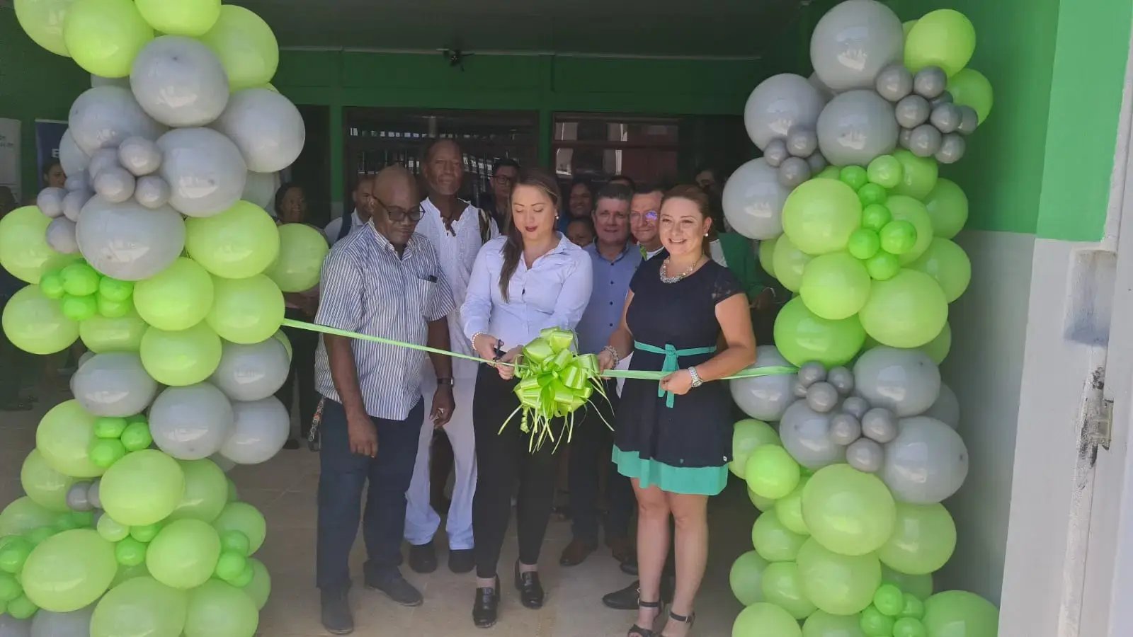 Personas participando en el corte de cinta durante la inauguración de un local decorado con globos verdes y grises.