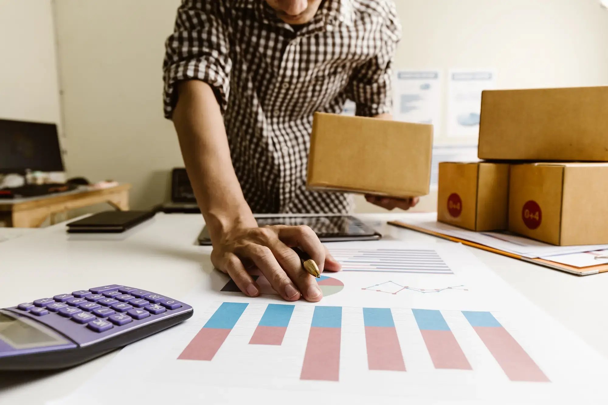 Hombre analizando gráficas financieras mientras sostiene una caja en su negocio de envíos.