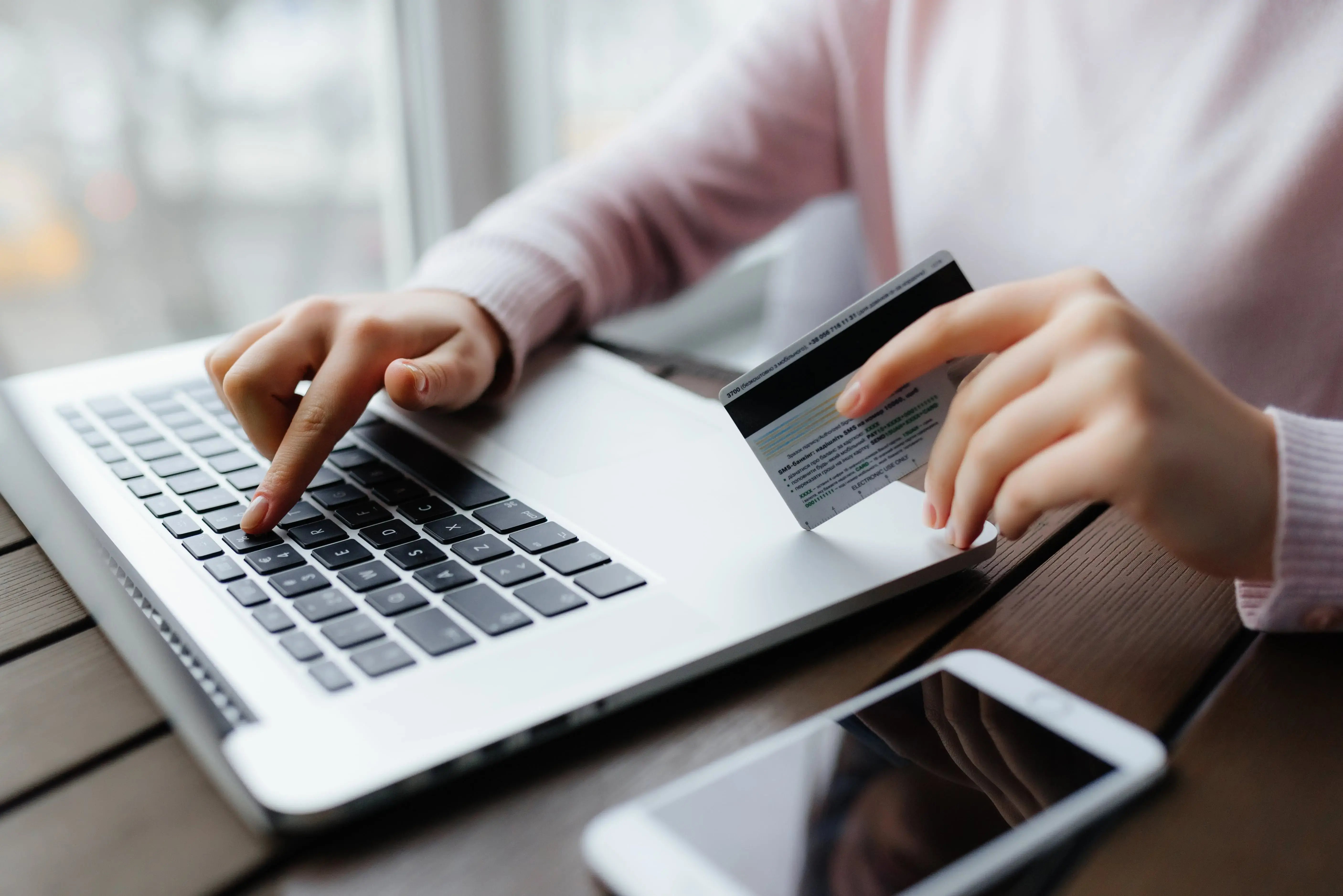 Persona usando una laptop con una tarjeta de crédito en la mano y un teléfono móvil sobre la mesa.