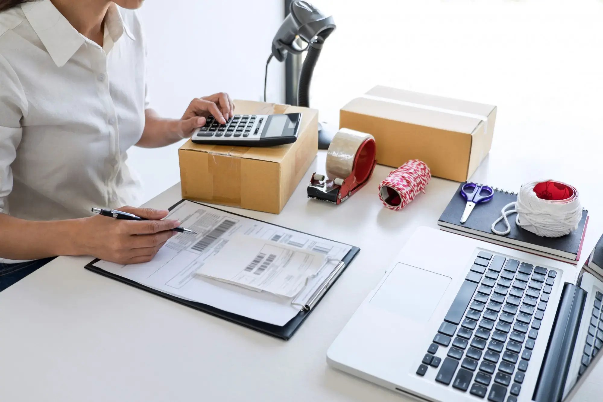 Mujer usando calculadora y revisando facturas junto a cajas de productos y laptop en escritorio.