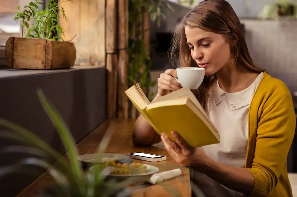 Mujer joven leyendo un libro amarillo y tomando café en una cafetería acogedora