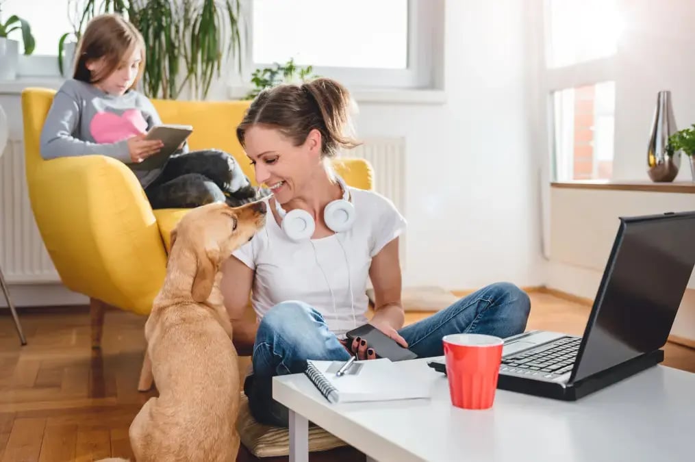 Mujer trabajando en casa con su laptop mientras acaricia a su perro y su hija usa una tableta.