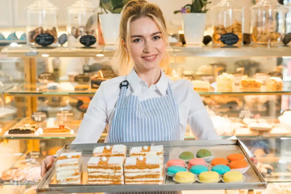 Mujer pastelera mostrando bandeja con pasteles y macarons en vitrina de panadería.