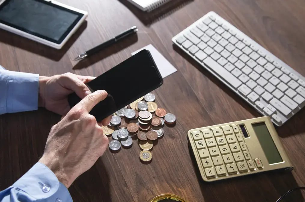 Persona usando un teléfono móvil sobre una mesa con monedas, calculadora y teclado.