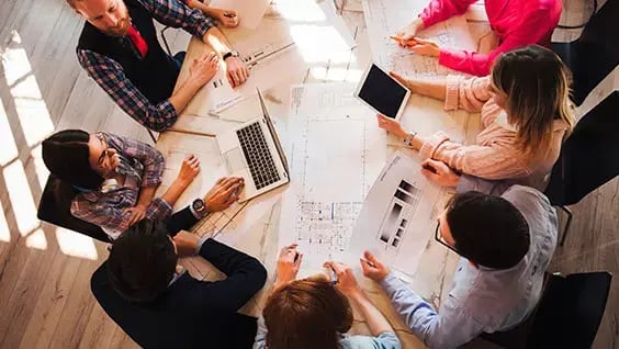 Grupo de personas reunidas alrededor de una mesa con planos, computadoras y tabletas en una sesión de trabajo colaborativo. Grupo de personas reunidas alrededor de una mesa con planos, computadoras y tabletas en una sesión de trabajo colaborativo.