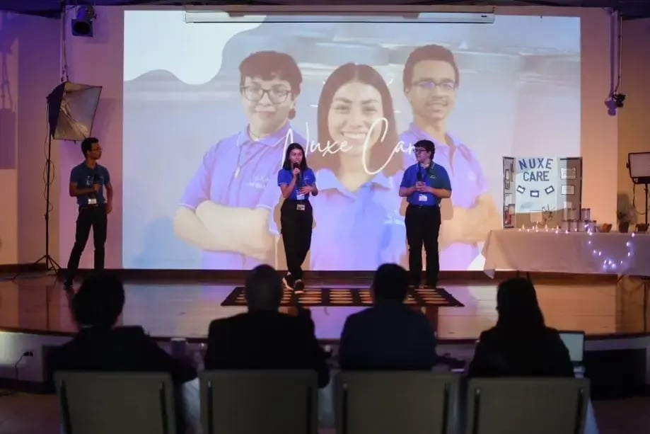Jóvenes exponiendo el proyecto Nuxe Care en un escenario con proyección de su equipo en pantalla.
