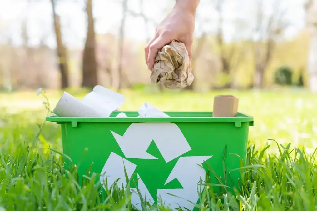 Mano depositando papel arrugado en un contenedor verde de reciclaje en un parque.