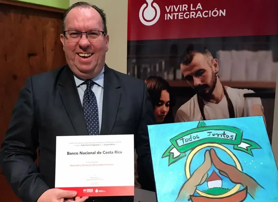 Hombre sonriente sostiene un reconocimiento otorgado al Banco Nacional de Costa Rica, acompañado de una pintura con la frase "Todos Juntos", en un evento sobre integración.