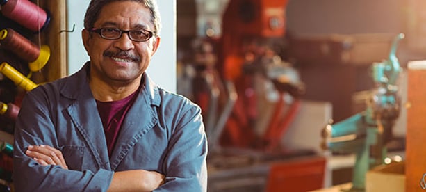 Hombre con bata de trabajo en un taller, cruzado de brazos y sonriendo, simbolizando emprendimiento o negocio propio.