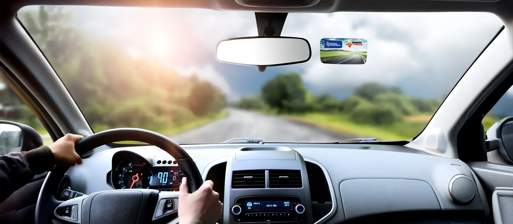 Vista en primera persona, manejando un auto con las manos en el volante sobre la carretera rodeada de vegetación y el sol entre nubes.