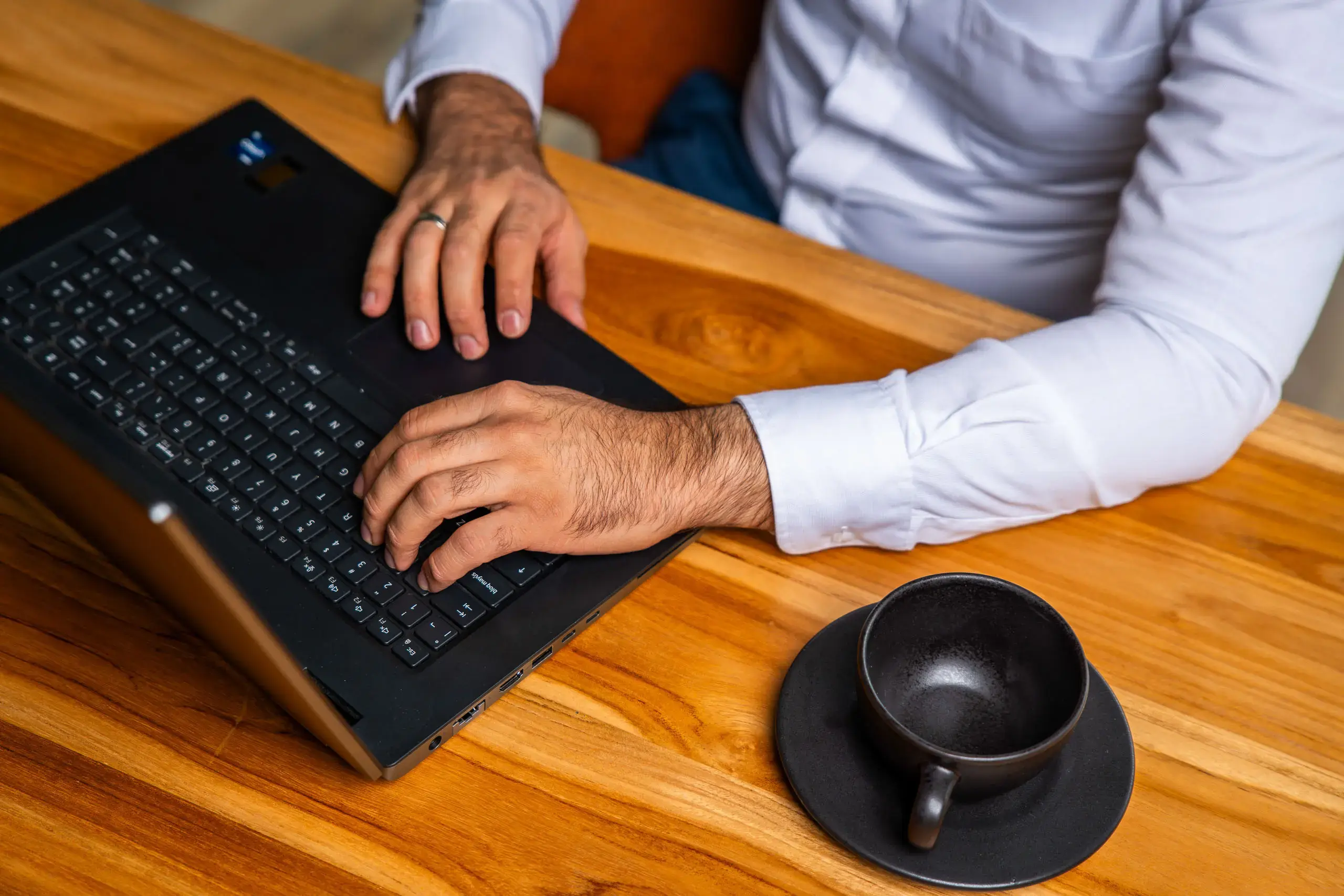Manos de un hombre escribiendo en una computadora portátil sobre una mesa de madera junto a una taza vacía.