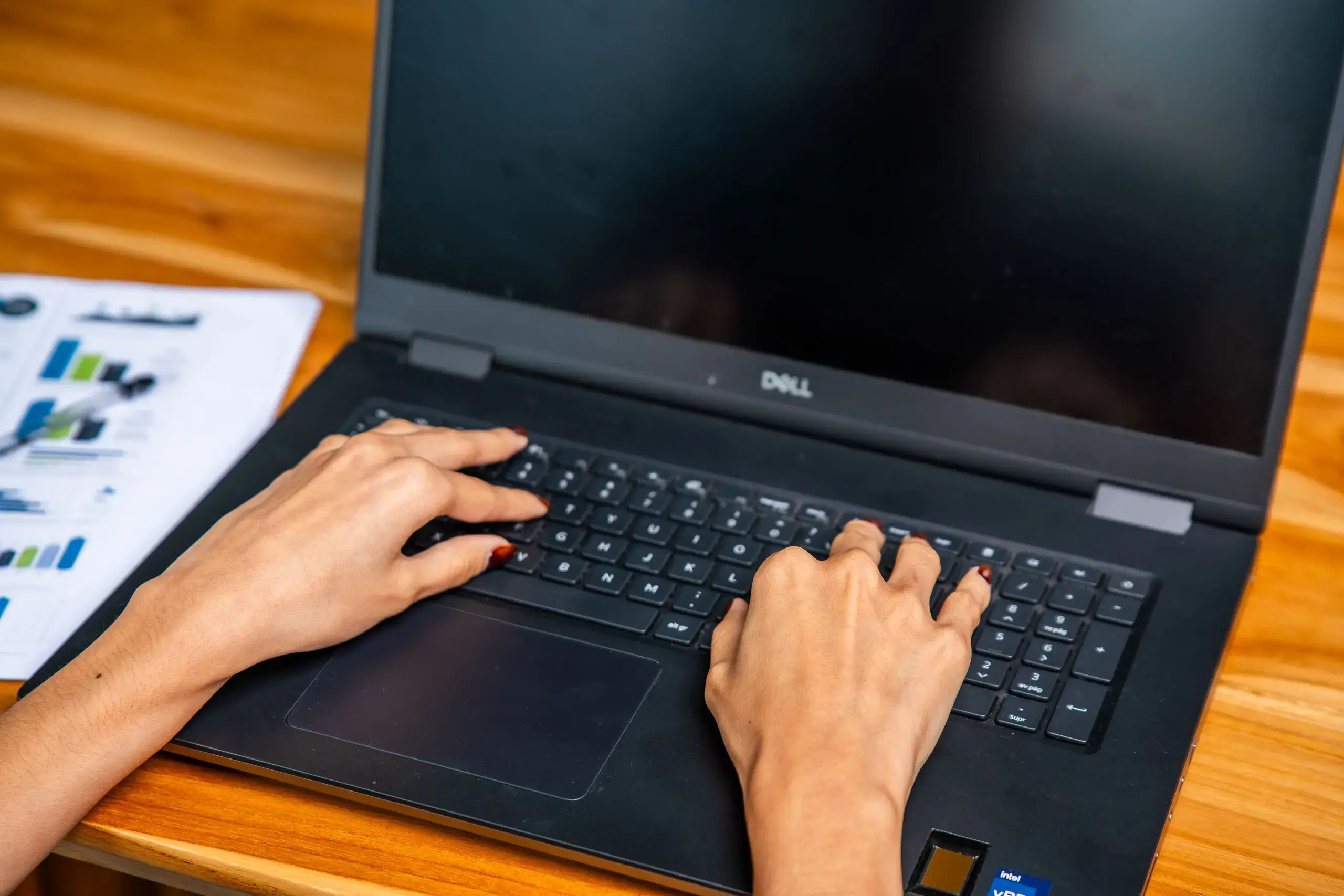 Manos escribiendo en un teclado de computadora portátil sobre un escritorio de madera con documentos a un lado.