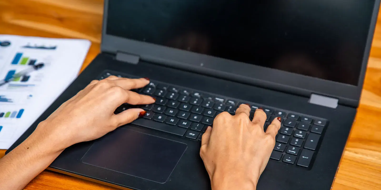 Manos escribiendo en un teclado de computadora portátil sobre un escritorio de madera con documentos a un lado.