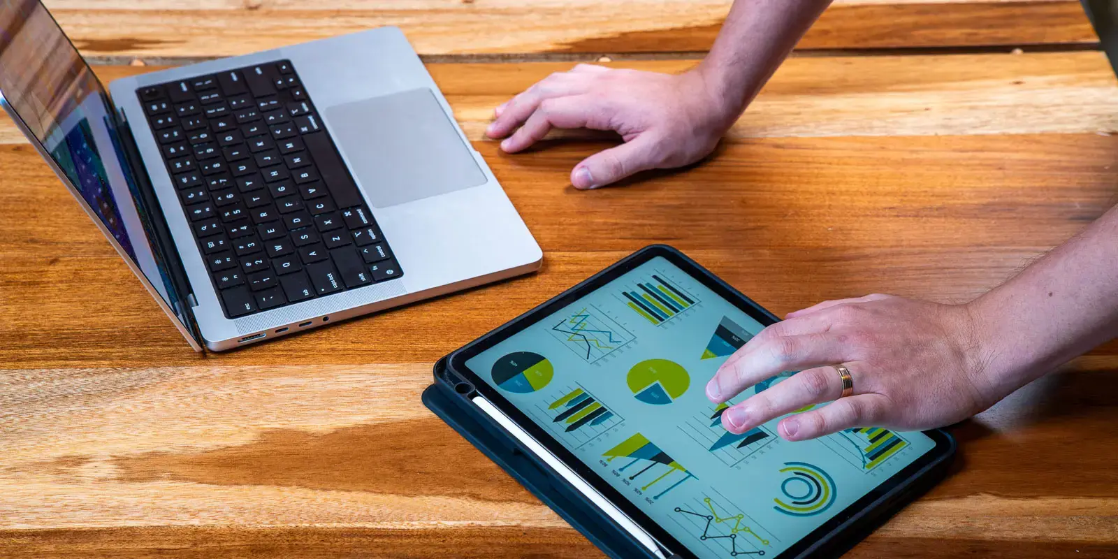 Mano interactuando con gráficos en una tablet junto a una computadora portátil sobre mesa de madera.