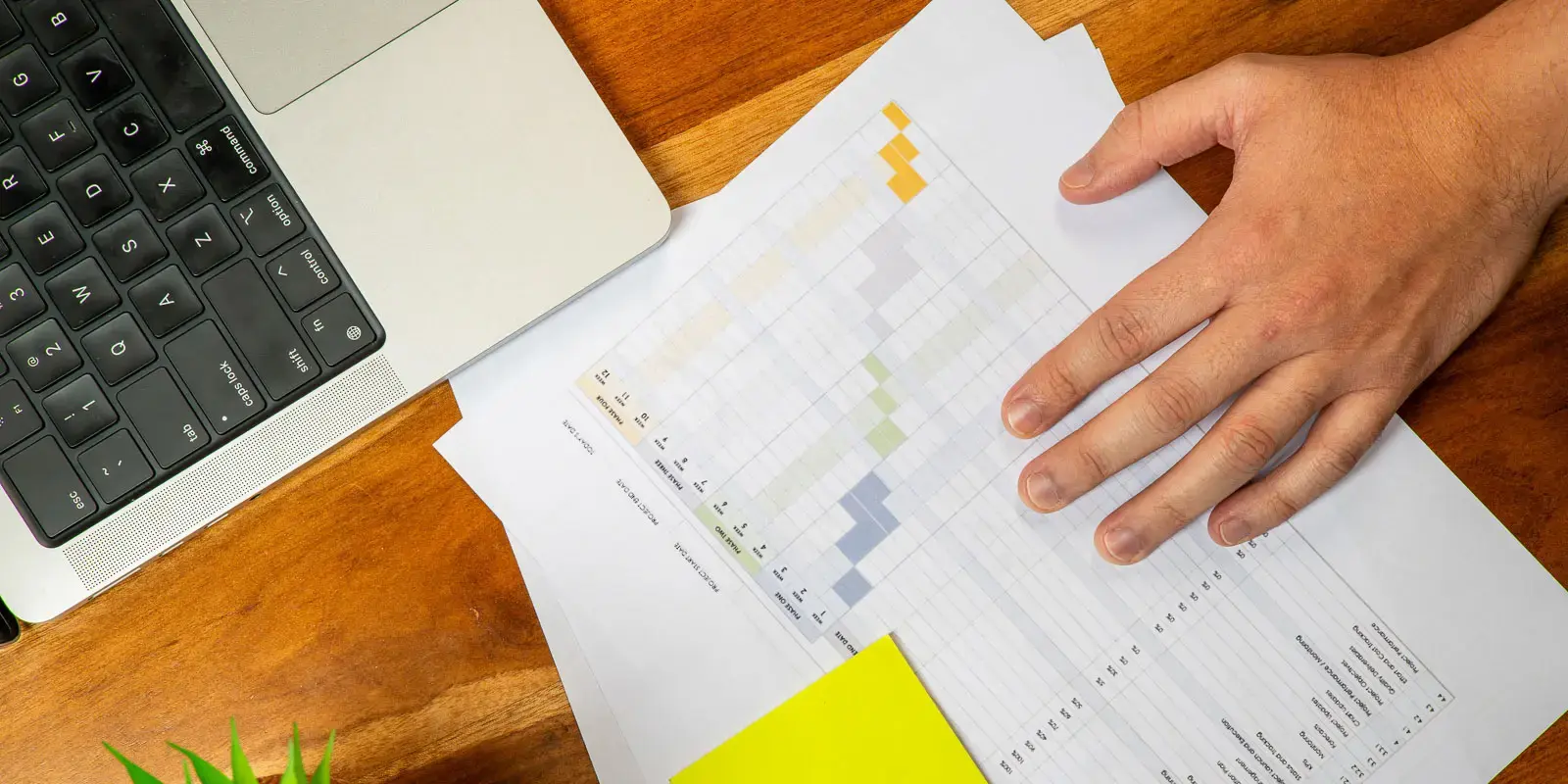 Mano sobre una hoja con tabla de datos junto a una computadora portátil sobre una mesa de madera.