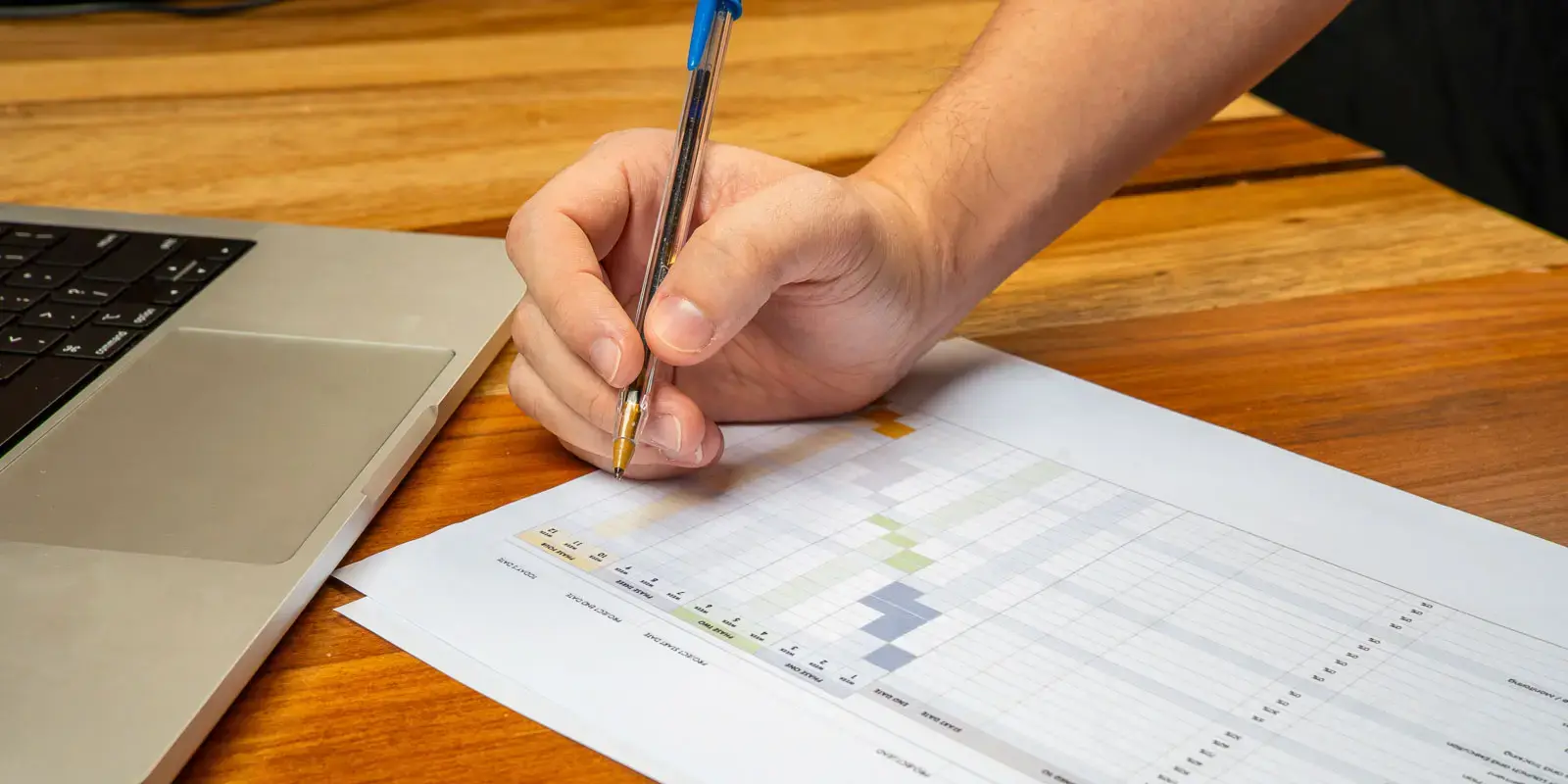 Mano rellenando una hoja de cálculo impresa junto a una laptop sobre una mesa de madera.