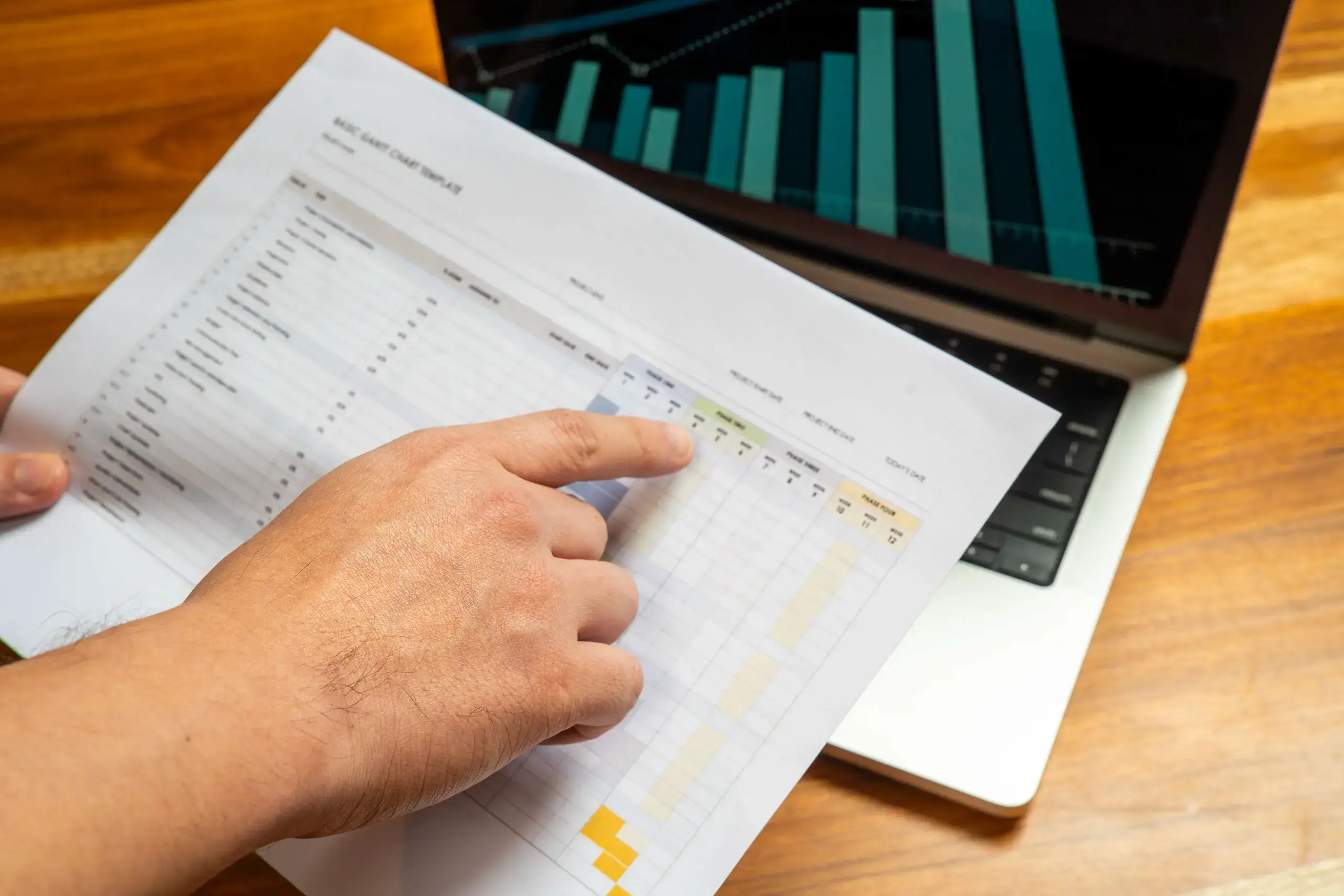 Mano señalando una hoja con tablas frente a una laptop que muestra gráficas.