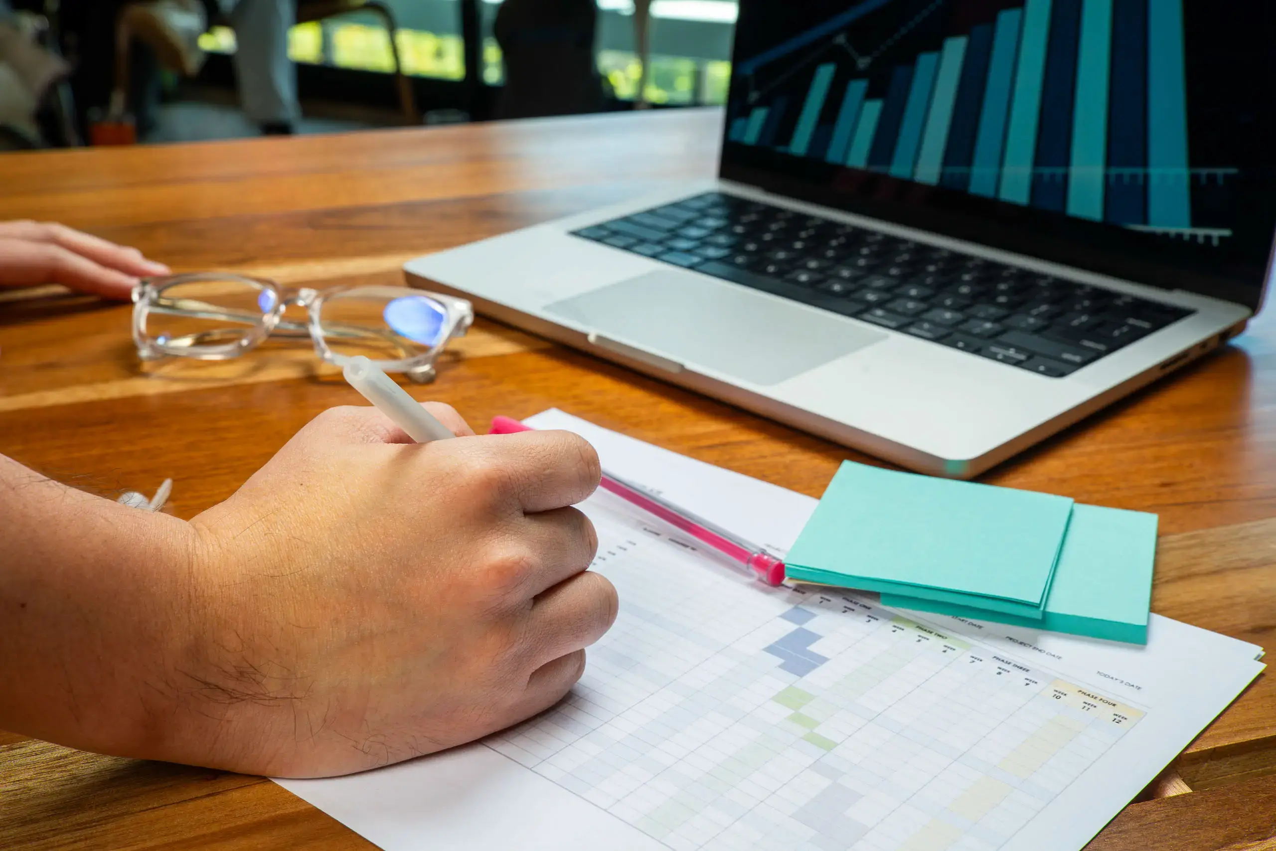 Mano escribiendo sobre una hoja con tablas mientras una laptop con gráficas está encendida sobre la mesa.