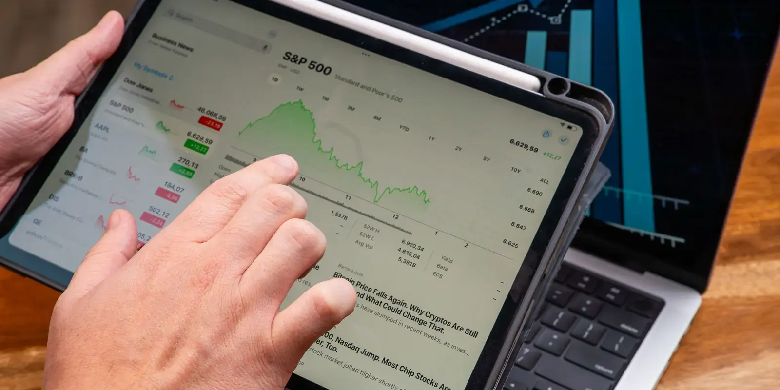Manos sosteniendo una tablet que muestra gráficos y datos del S&P 500 sobre un escritorio con una laptop al fondo.