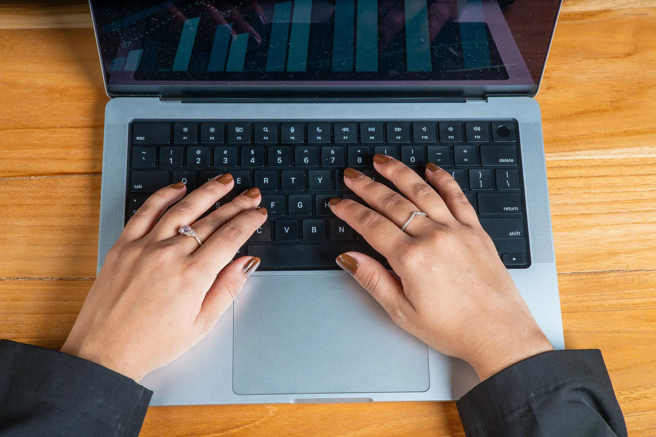 Manos con anillos y uñas esmaltadas escribiendo en el teclado de una laptop sobre una superficie de madera.