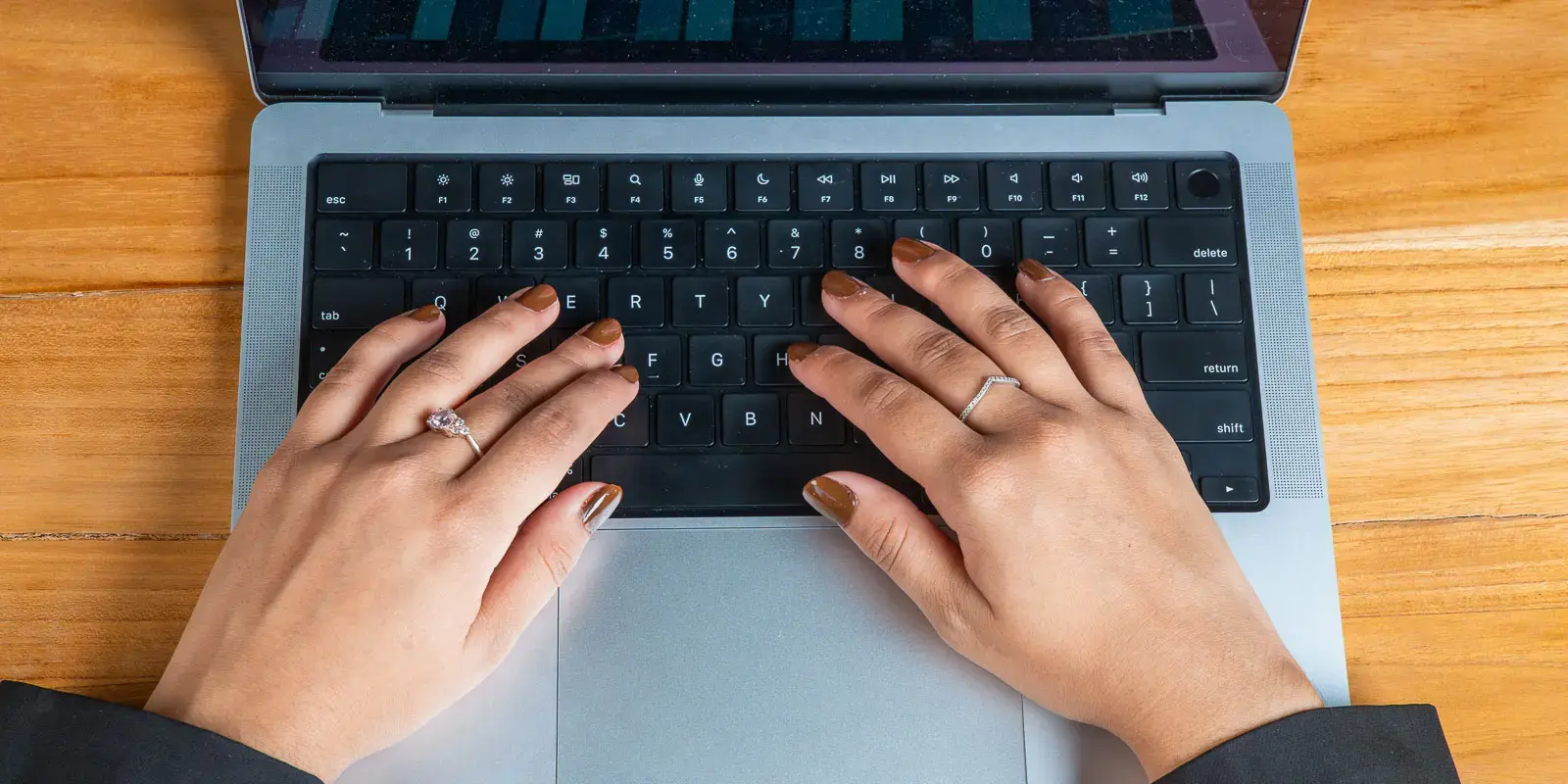 Manos con anillos y uñas esmaltadas escribiendo en el teclado de una laptop sobre una superficie de madera.