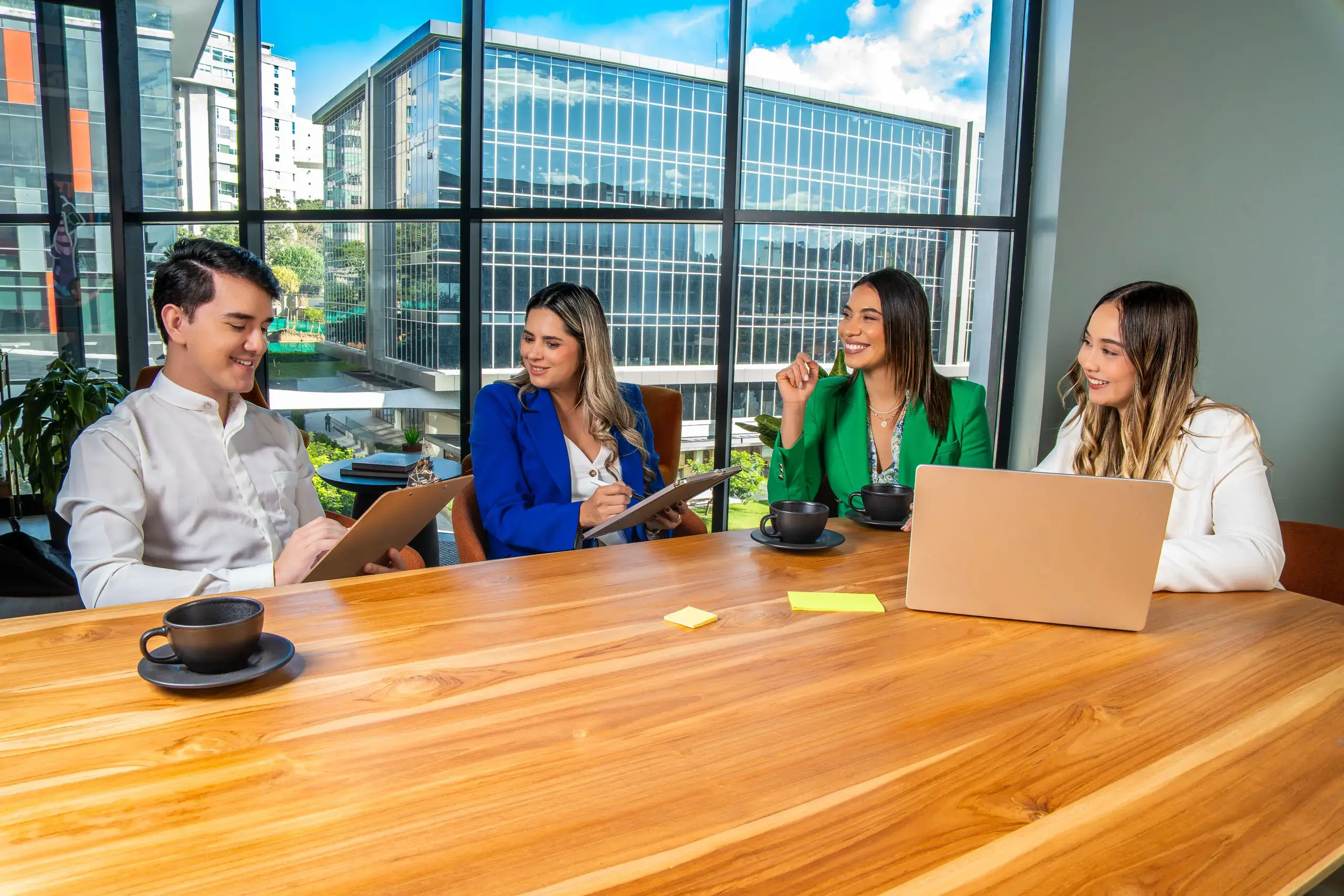 Grupo de cuatro personas conversando en una sala de reuniones con mesa de madera y vista a edificios acristalados.