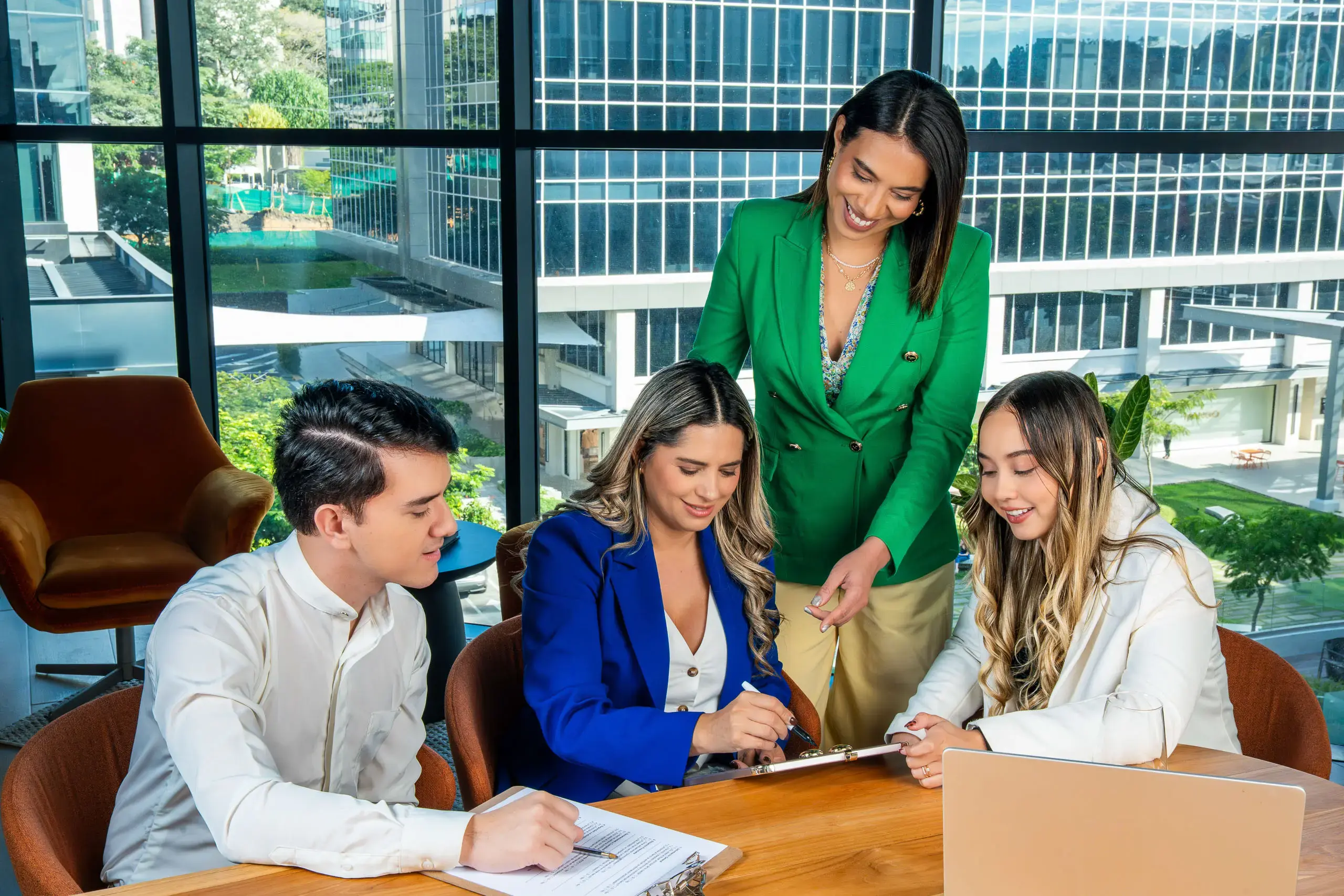 Grupo de cuatro personas reunidas en una mesa de oficina, colaborando y revisando documentos frente a una ventana con vista a edificios.